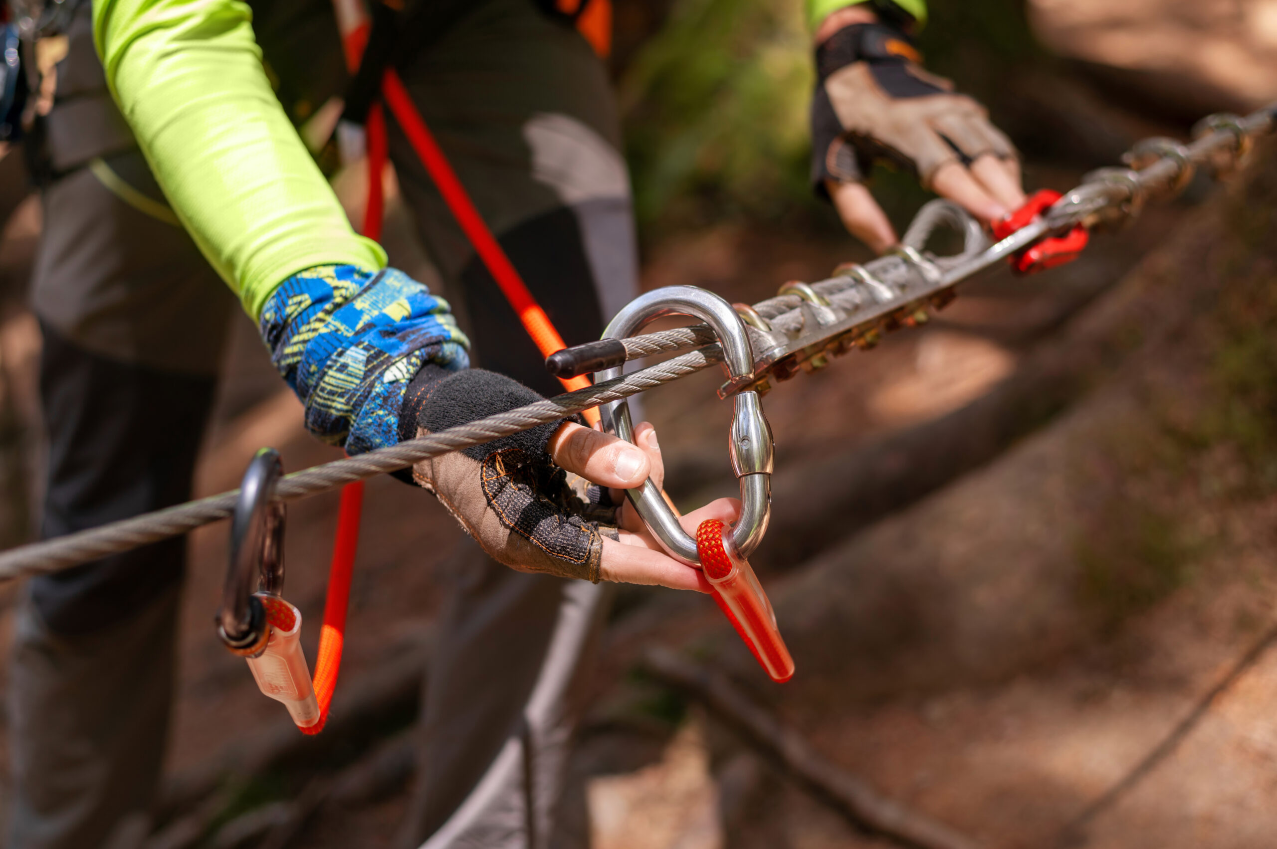 A close-up of a person's gloved hands holding a rappelling device and attaching it to their harness using a carabiner.